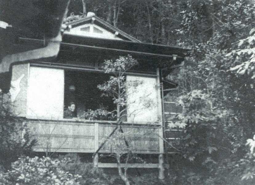 木立に囲まれた雨の念佛荘の外観。縁側の障子越しに人影が見える、歴史的写真。